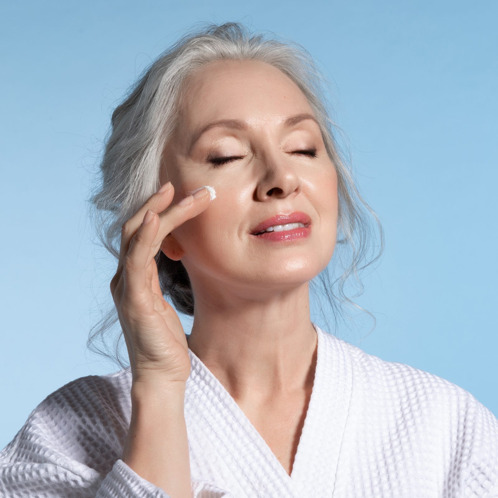 Senior woman with eyes closed putting cosmetic moisturizing rejuvenating cream on face feeling pleasure. Studio portrait on blue. Refreshment and anti-wrinkle skin therapy