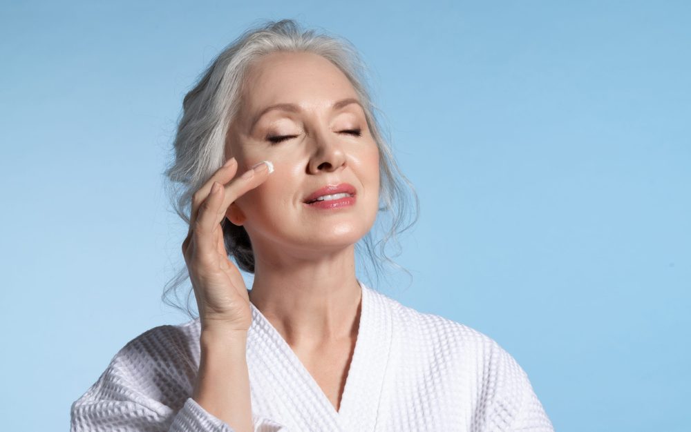 Senior woman with eyes closed putting cosmetic moisturizing rejuvenating cream on face feeling pleasure. Studio portrait on blue. Refreshment and anti-wrinkle skin therapy