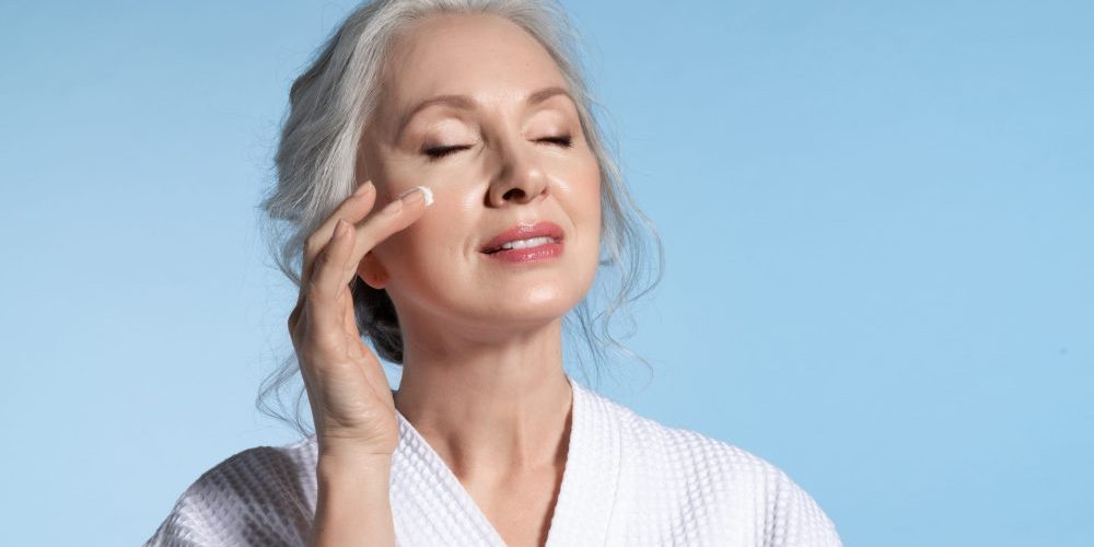 Senior woman with eyes closed putting cosmetic moisturizing rejuvenating cream on face feeling pleasure. Studio portrait on blue. Refreshment and anti-wrinkle skin therapy